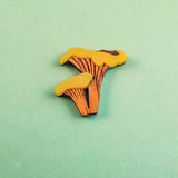 A small, flat illustration of two orange and yellow mushrooms lies on a light green surface—perfect for an eco-friendly Chanterelle Mushroom Pin or other handcrafted accessories.