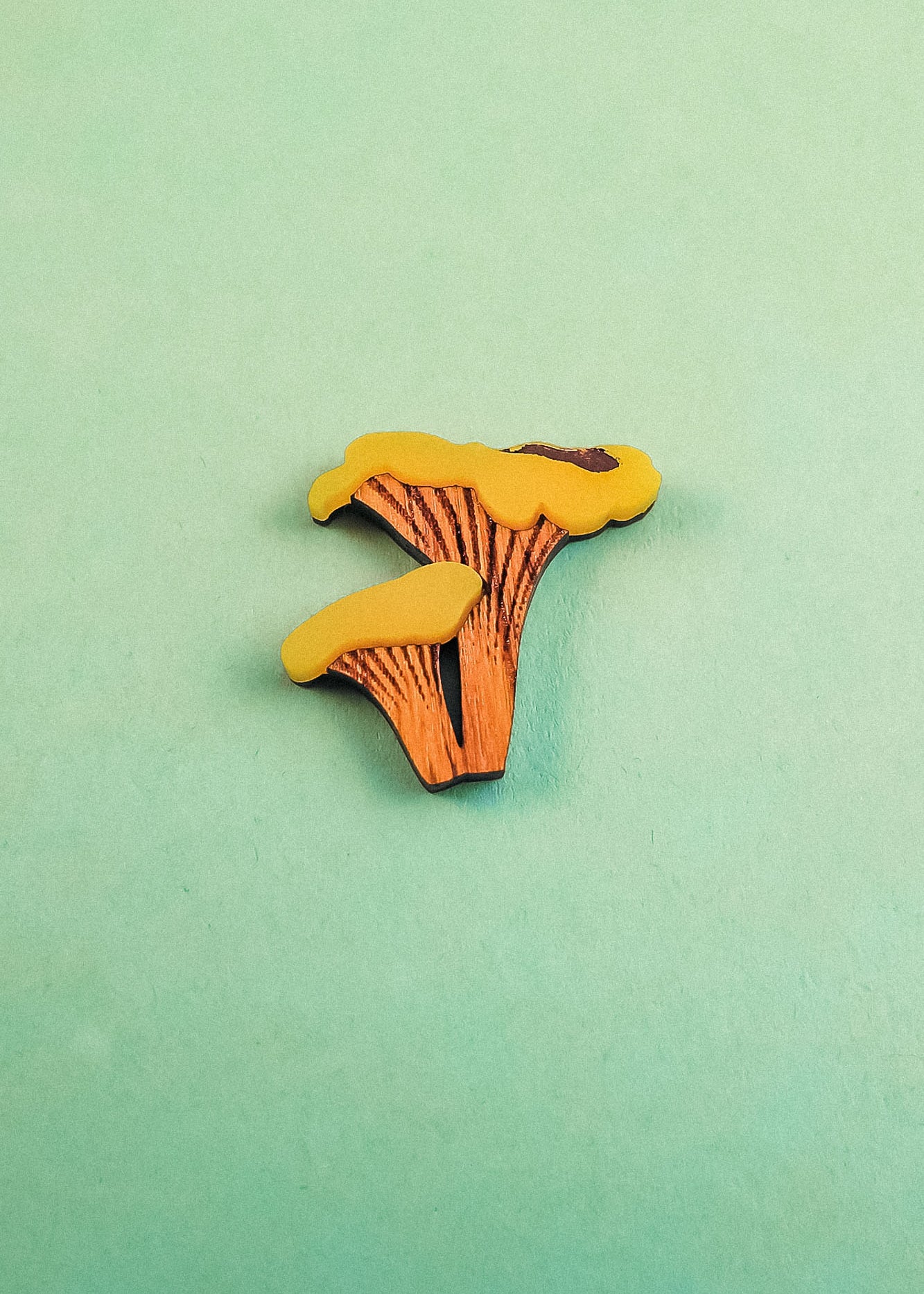 A small, flat illustration of two orange and yellow mushrooms lies on a light green surface—perfect for an eco-friendly Chanterelle Mushroom Pin or other handcrafted accessories.