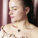 A woman with light skin and braided hair wears a cream off-shoulder top, a minimalistic gold circle necklace with matching earrings, posing in front of pink curtains.