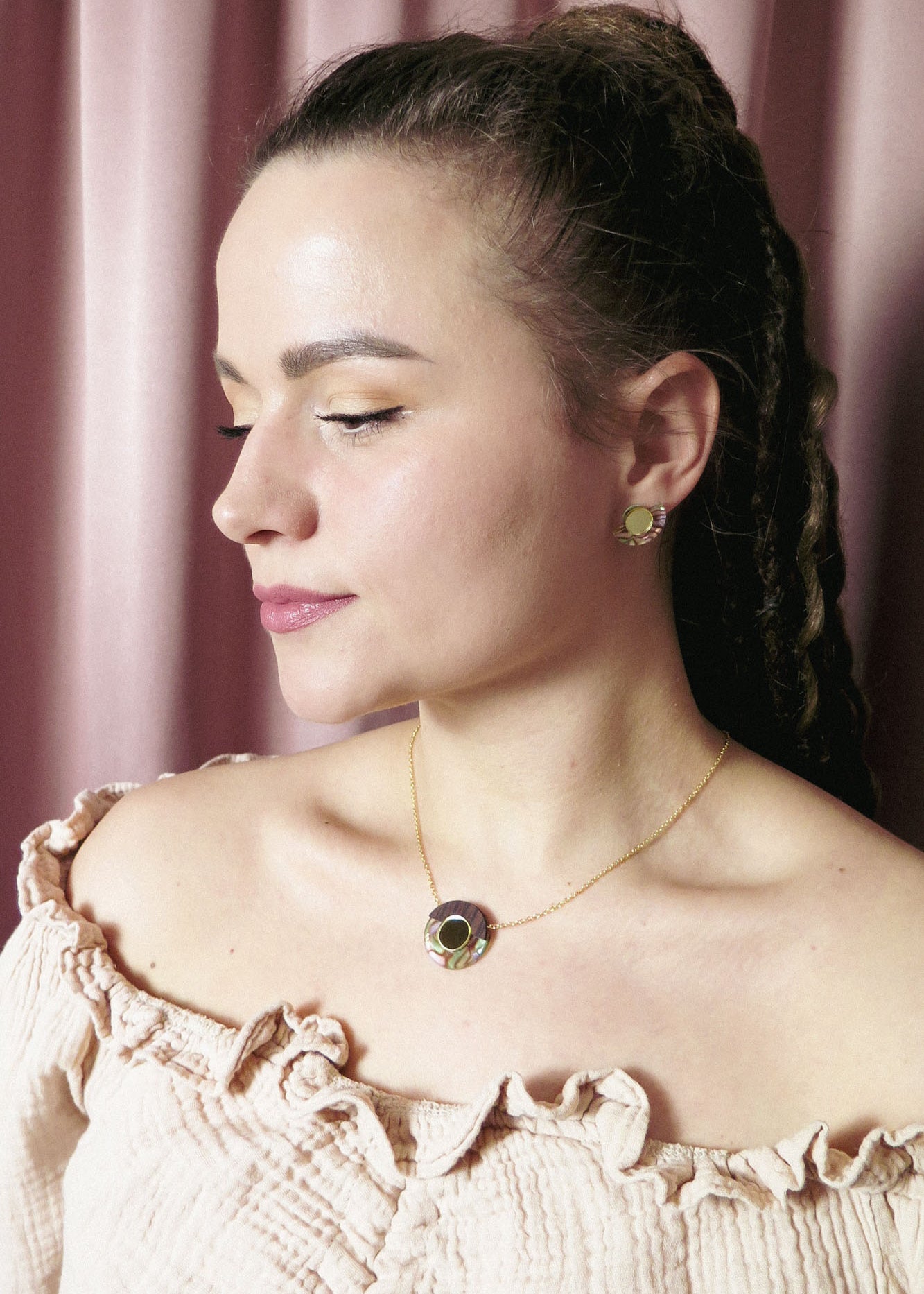 A woman with light skin and braided hair wears a cream off-shoulder top, a minimalistic gold circle necklace with matching earrings, posing in front of pink curtains.