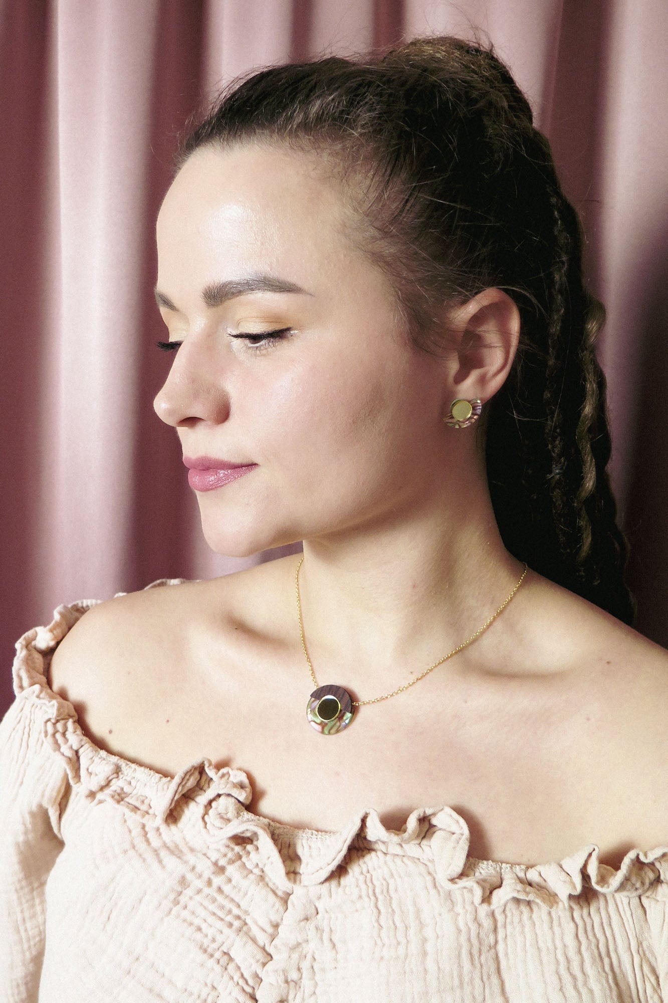 A woman with light skin and braided hair wears a cream off-shoulder top, a minimalistic gold circle necklace with matching earrings, posing in front of pink curtains.