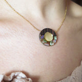 A close-up of a person wearing a minimalistic gold Circle Necklace with a round pendant featuring a gold circle surrounded by iridescent abalone shell details, against fair skin and a light textured top—perfect for any capsule wardrobe.