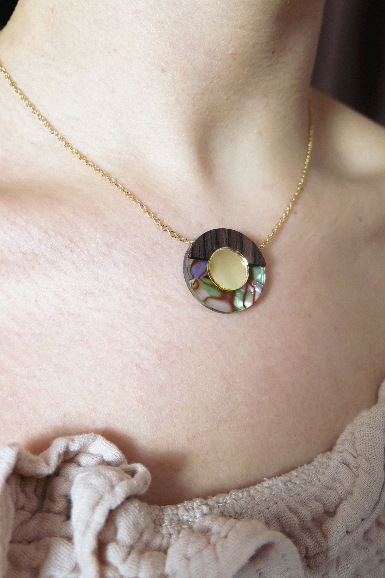 A close-up of a person wearing a minimalistic gold Circle Necklace with a round pendant featuring a gold circle surrounded by iridescent abalone shell details, against fair skin and a light textured top—perfect for any capsule wardrobe.