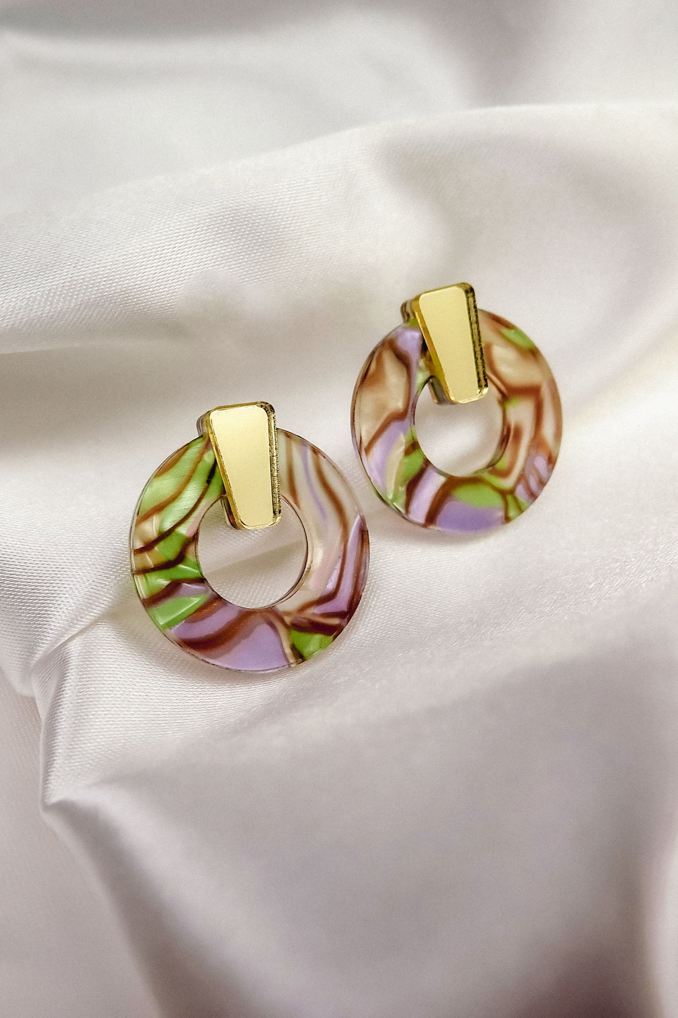 A pair of hypoallergenic Minimalist Circle Stud Earrings with gold rectangular tops and marbled acrylic glass circular bottoms in pink, green, and brown, displayed on white satin fabric.