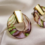 Close-up of a round, marbled earring made from acrylic glass with pink, green, and purple swirls, featuring a shiny gold geometric accent on top. This hypoallergenic piece rests elegantly on soft, cream-colored satin fabric.