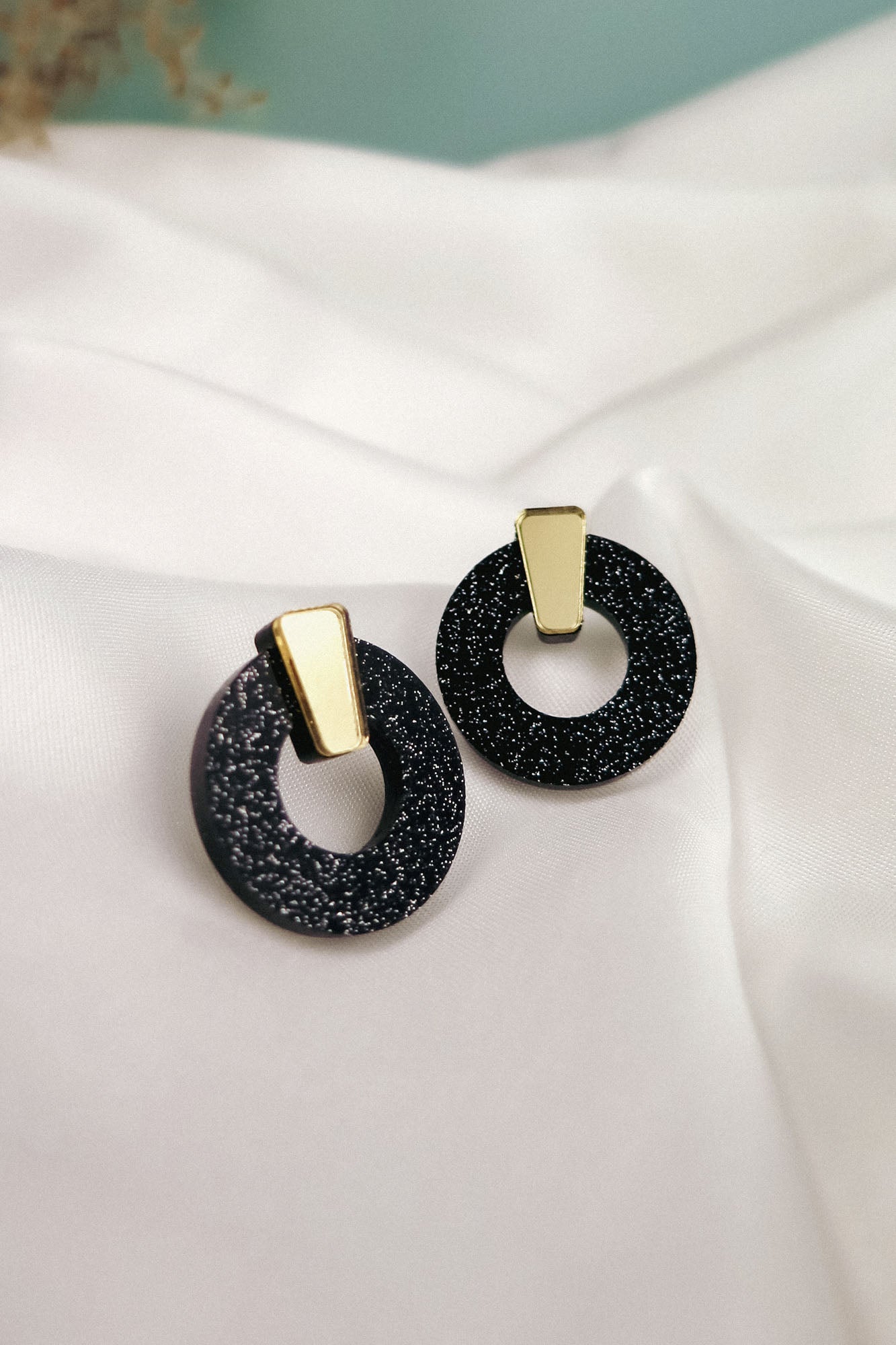A pair of stylish Minimalist Circle Stud Earrings featuring black glittery acrylic glass hoops with gold rectangular accents, displayed on a white satin fabric background. Hypoallergenic for comfortable, all-day wear.