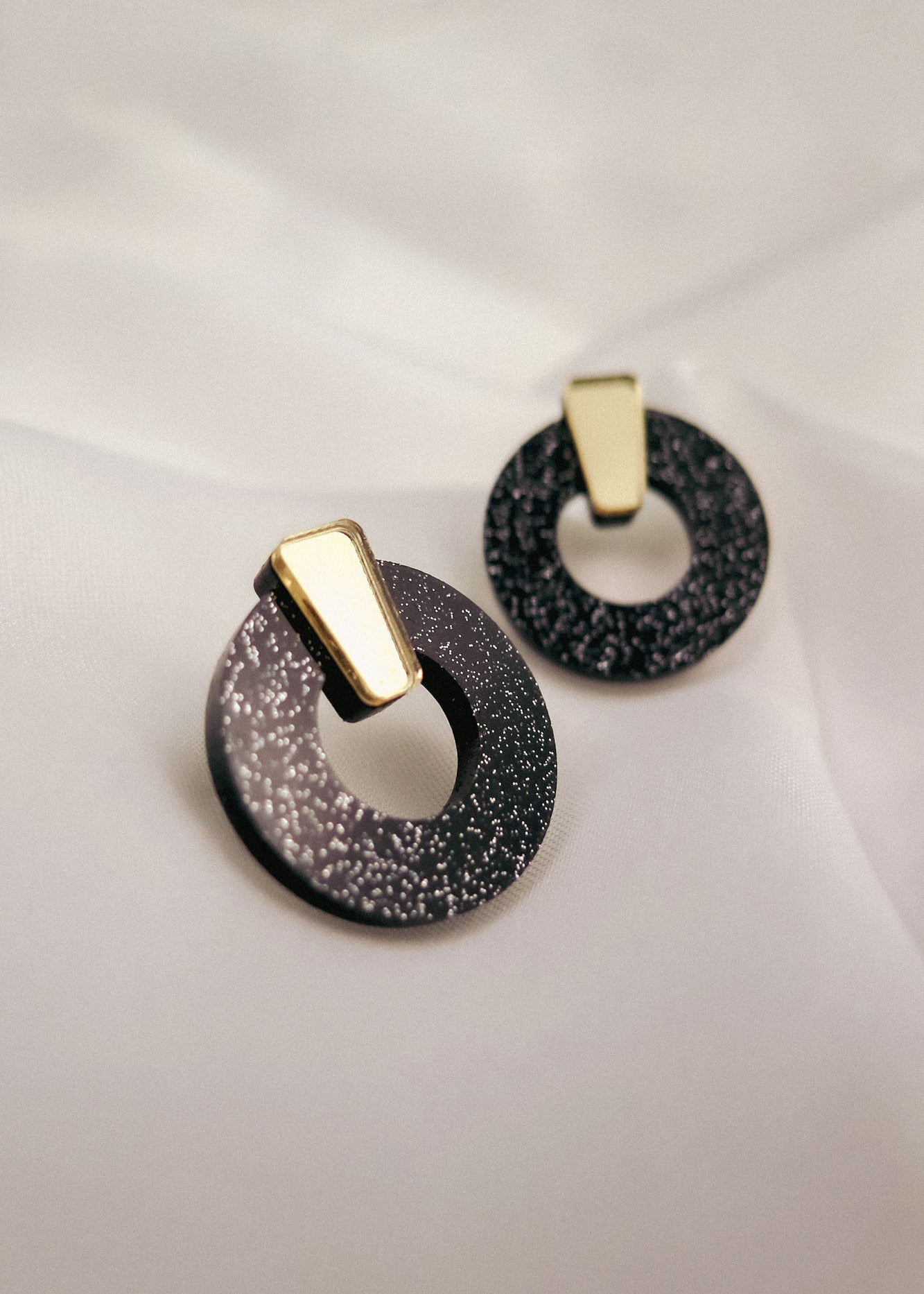 Minimalist Circle Stud Earrings crafted from hypoallergenic acrylic glass, these round black earrings feature silver speckles and gold geometric accents, displayed on a soft white fabric background.