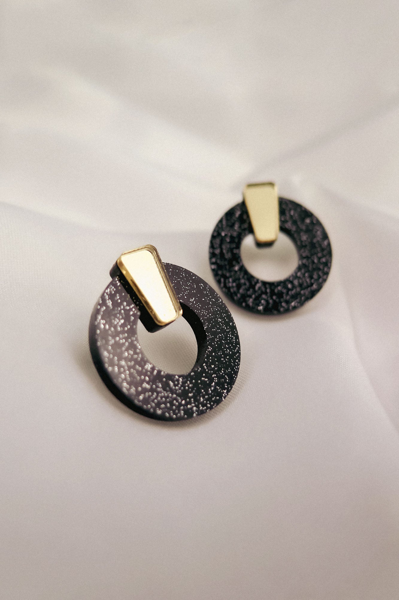 Minimalist Circle Stud Earrings crafted from hypoallergenic acrylic glass, these round black earrings feature silver speckles and gold geometric accents, displayed on a soft white fabric background.