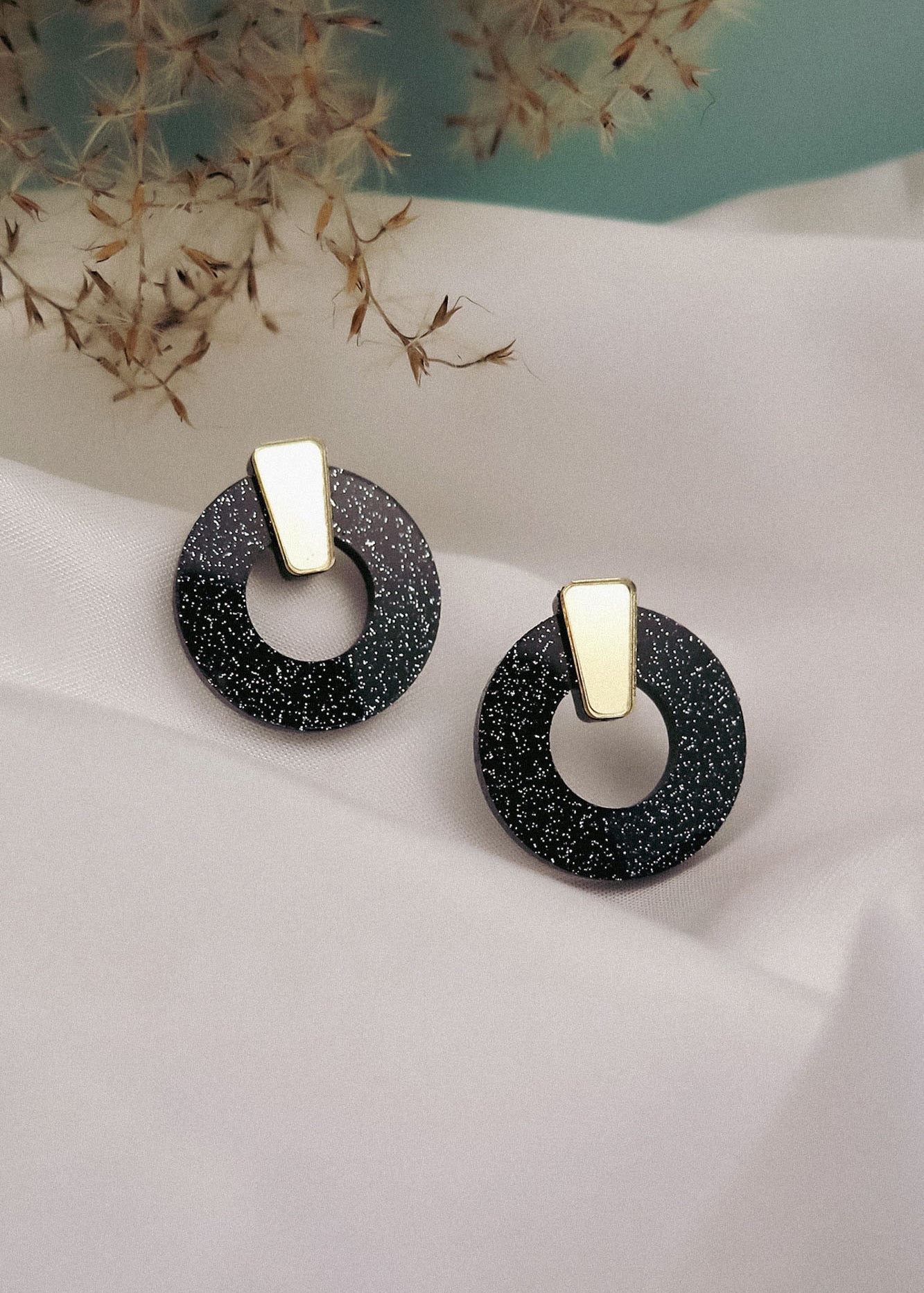 A pair of minimalist circle stud earrings in black acrylic glass with white speckles and gold triangular studs, displayed on white fabric with dried grasses in the background. Hypoallergenic for comfortable, everyday wear.