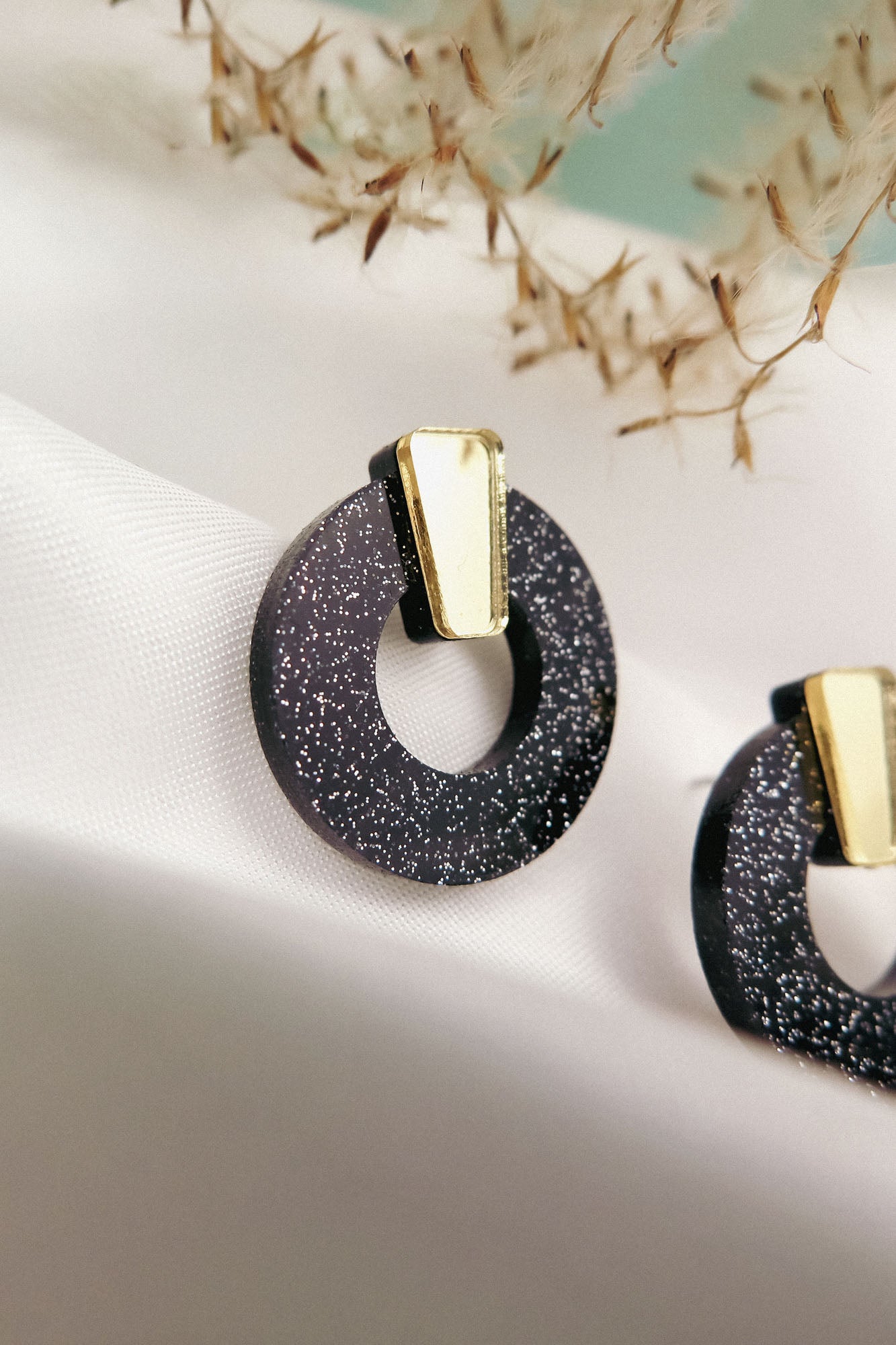 Close-up of minimalist circle stud earrings in black acrylic glass with silver glitter and a gold accent, displayed on a white fabric background with blurred dried plant details above. Hypoallergenic design ensures comfortable wear.