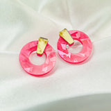 A pair of Minimalist Circle Stud Earrings featuring large, faceted pink acrylic glass hoops and small, irregular yellow rectangular studs, displayed on a soft white satin fabric background.