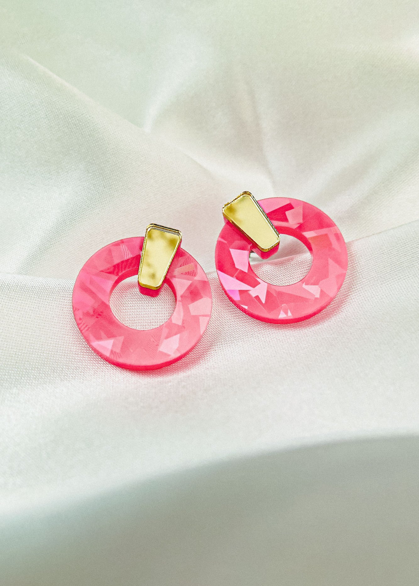 A pair of Minimalist Circle Stud Earrings featuring large, faceted pink acrylic glass hoops and small, irregular yellow rectangular studs, displayed on a soft white satin fabric background.
