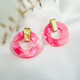 A pair of Minimalist Circle Stud Earrings featuring shiny pink acrylic glass pendants and gold accents, displayed on a white fabric background with blurred dried flowers in the corner.