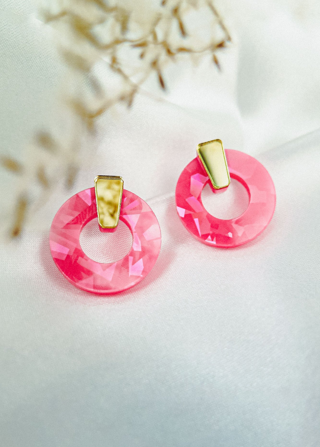 A pair of Minimalist Circle Stud Earrings featuring shiny pink acrylic glass pendants and gold accents, displayed on a white fabric background with blurred dried flowers in the corner.