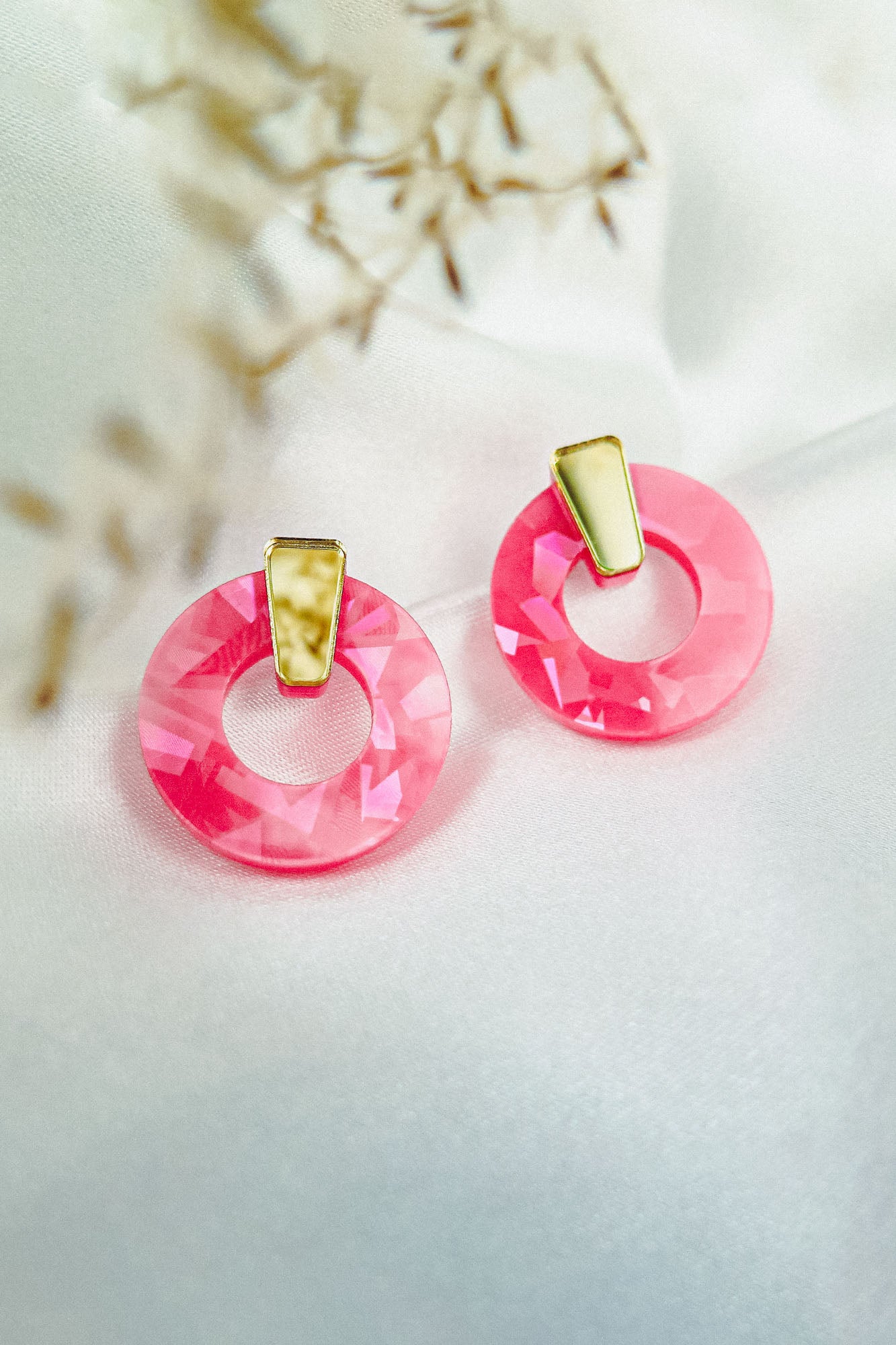 A pair of Minimalist Circle Stud Earrings featuring shiny pink acrylic glass pendants and gold accents, displayed on a white fabric background with blurred dried flowers in the corner.