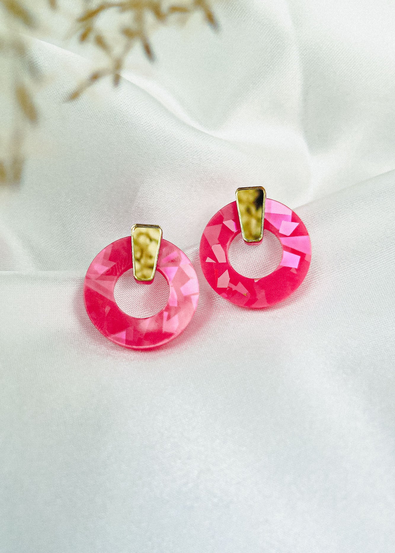 A pair of Minimalist Circle Stud Earrings with round, faceted pink acrylic glass and gold-tone accents rest on a smooth white fabric background. Delicate, out-of-focus dried flowers appear in the top left corner.