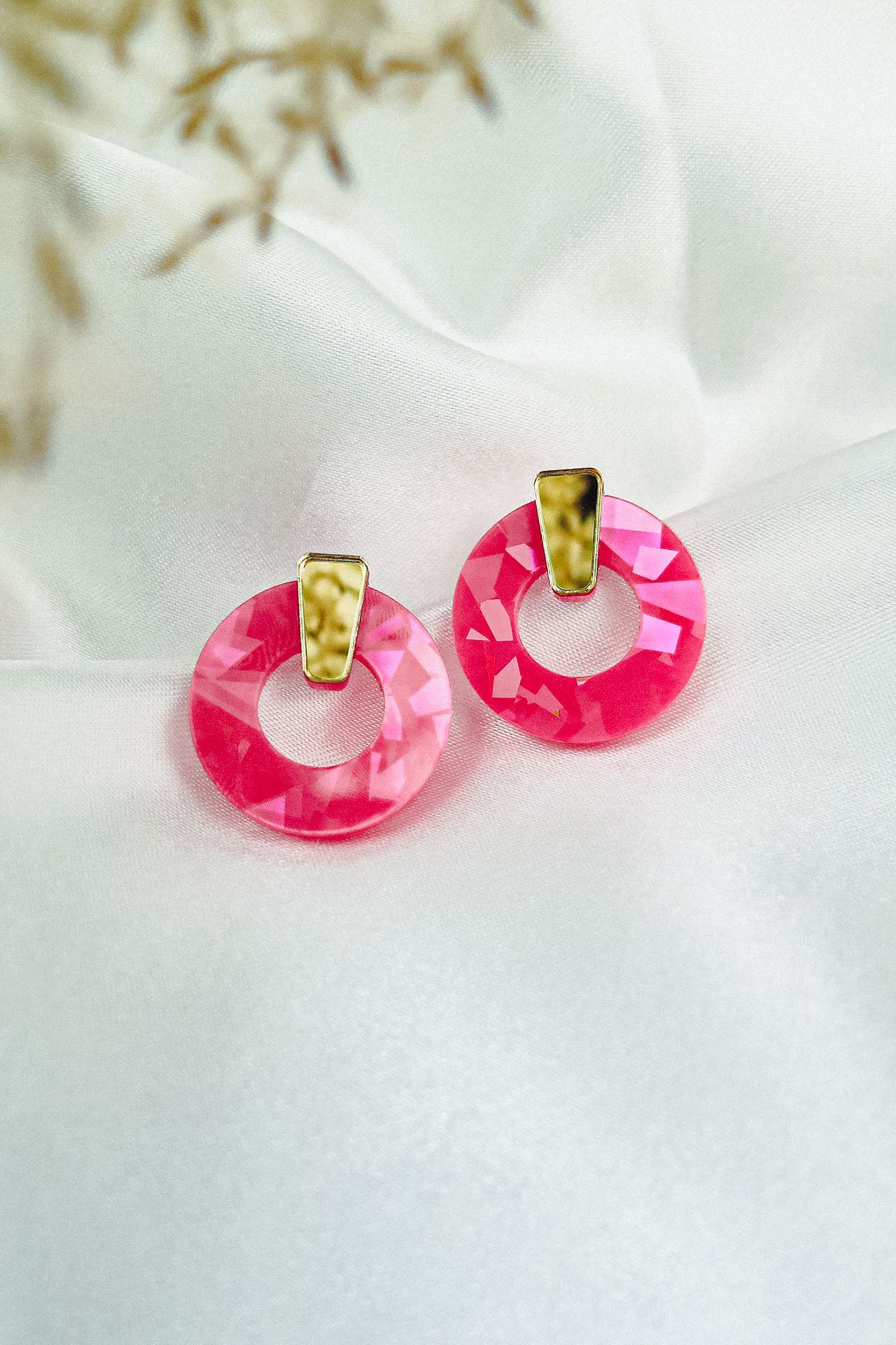 A pair of Minimalist Circle Stud Earrings with round, faceted pink acrylic glass and gold-tone accents rest on a smooth white fabric background. Delicate, out-of-focus dried flowers appear in the top left corner.