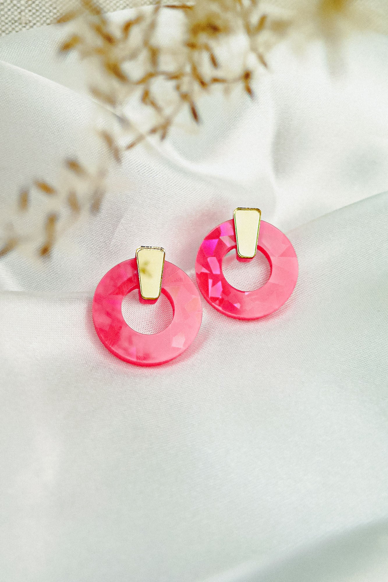 A pair of minimalist circle stud earrings with faceted pink acrylic glass and subtle gold accents, displayed on white satin fabric with soft, blurred dried plant accents in the foreground.