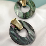 Minimalist Circle Stud Earrings crafted from acrylic glass feature marbled green, black, and purple hues with gold rectangular accents. They rest on white textured fabric, against a softly blurred light blue backdrop. Hypoallergenic for sensitive ears.