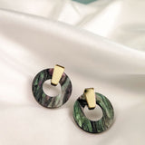 A pair of hypoallergenic Minimalist Circle Stud Earrings, featuring round marbled green acrylic glass and gold rectangular studs, are displayed on a white satin fabric background.
