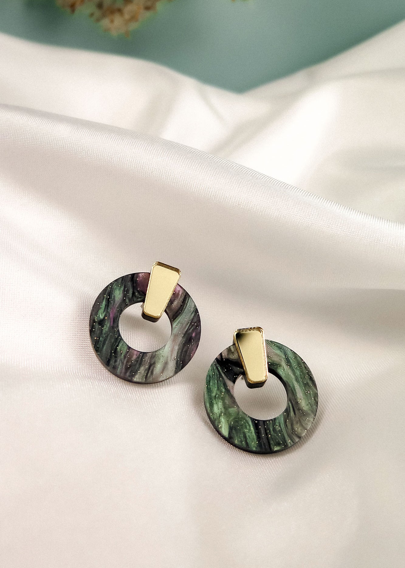 A pair of hypoallergenic Minimalist Circle Stud Earrings, featuring round marbled green acrylic glass and gold rectangular studs, are displayed on a white satin fabric background.