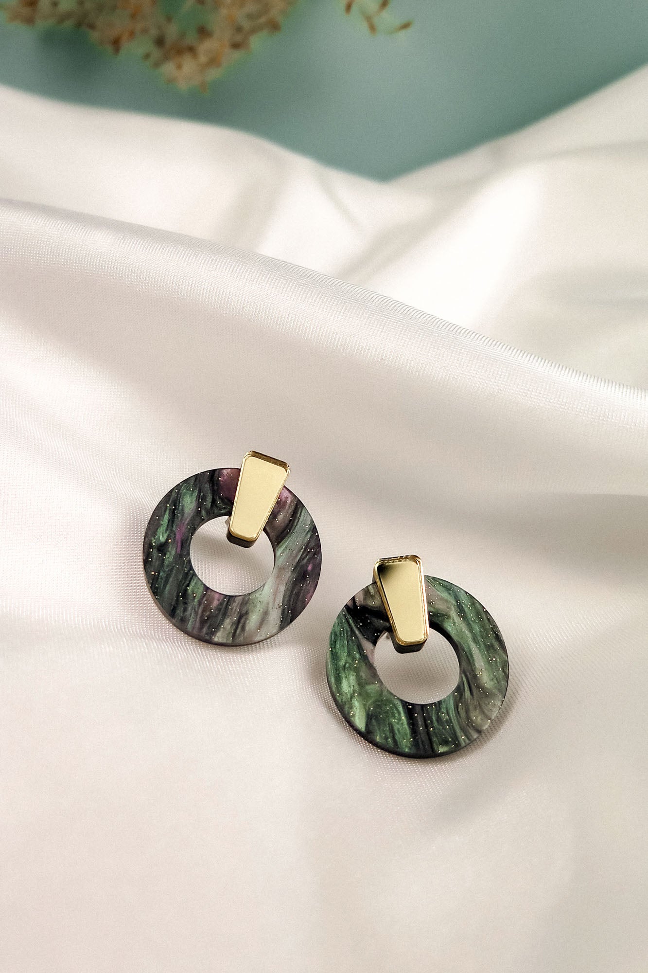 A pair of hypoallergenic Minimalist Circle Stud Earrings, featuring round marbled green acrylic glass and gold rectangular studs, are displayed on a white satin fabric background.