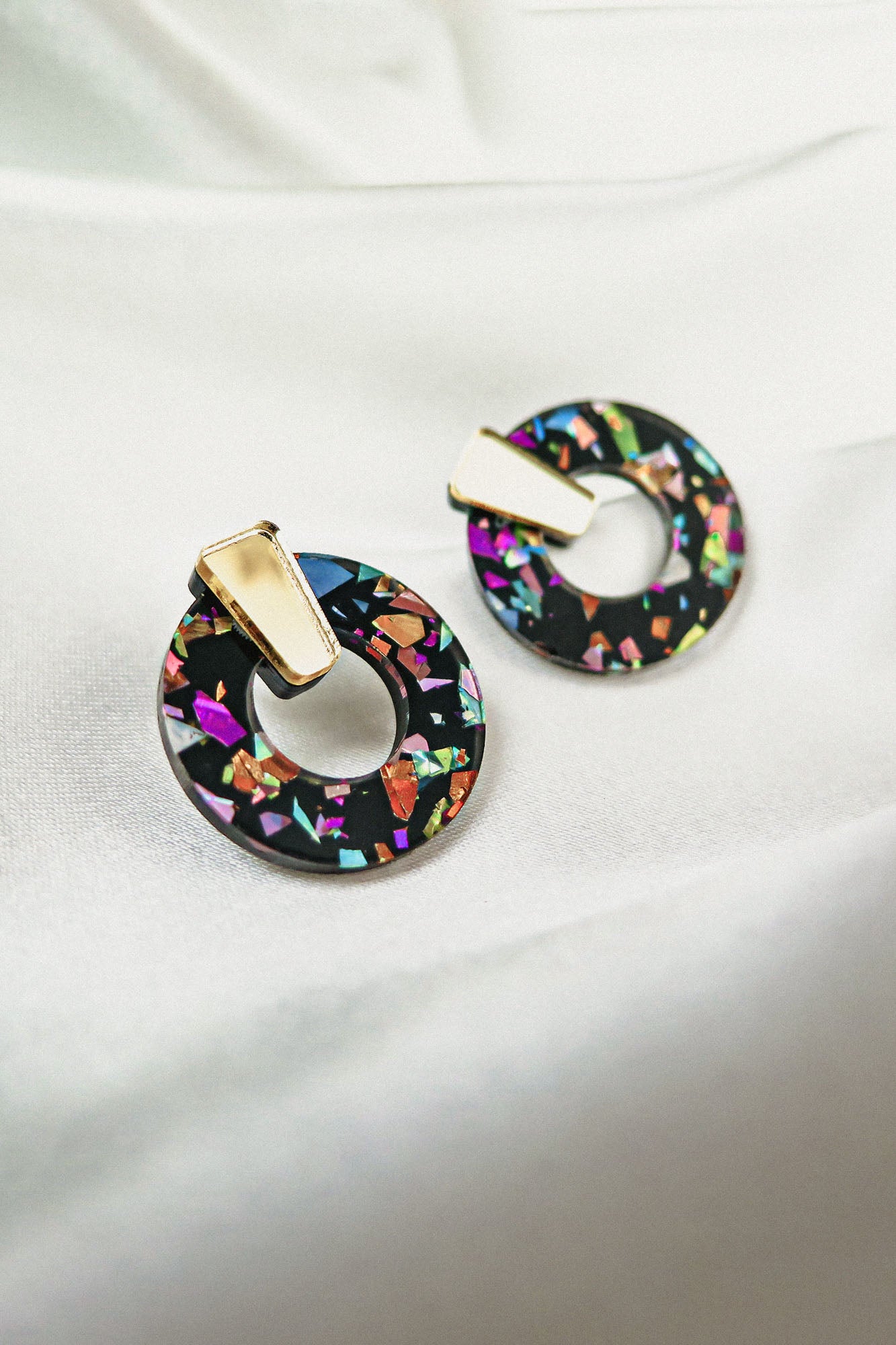 A pair of Minimalist Circle Stud Earrings made from hypoallergenic acrylic glass, featuring a black base with colorful, iridescent shards and gold-toned rectangular studs, displayed on a white fabric background.