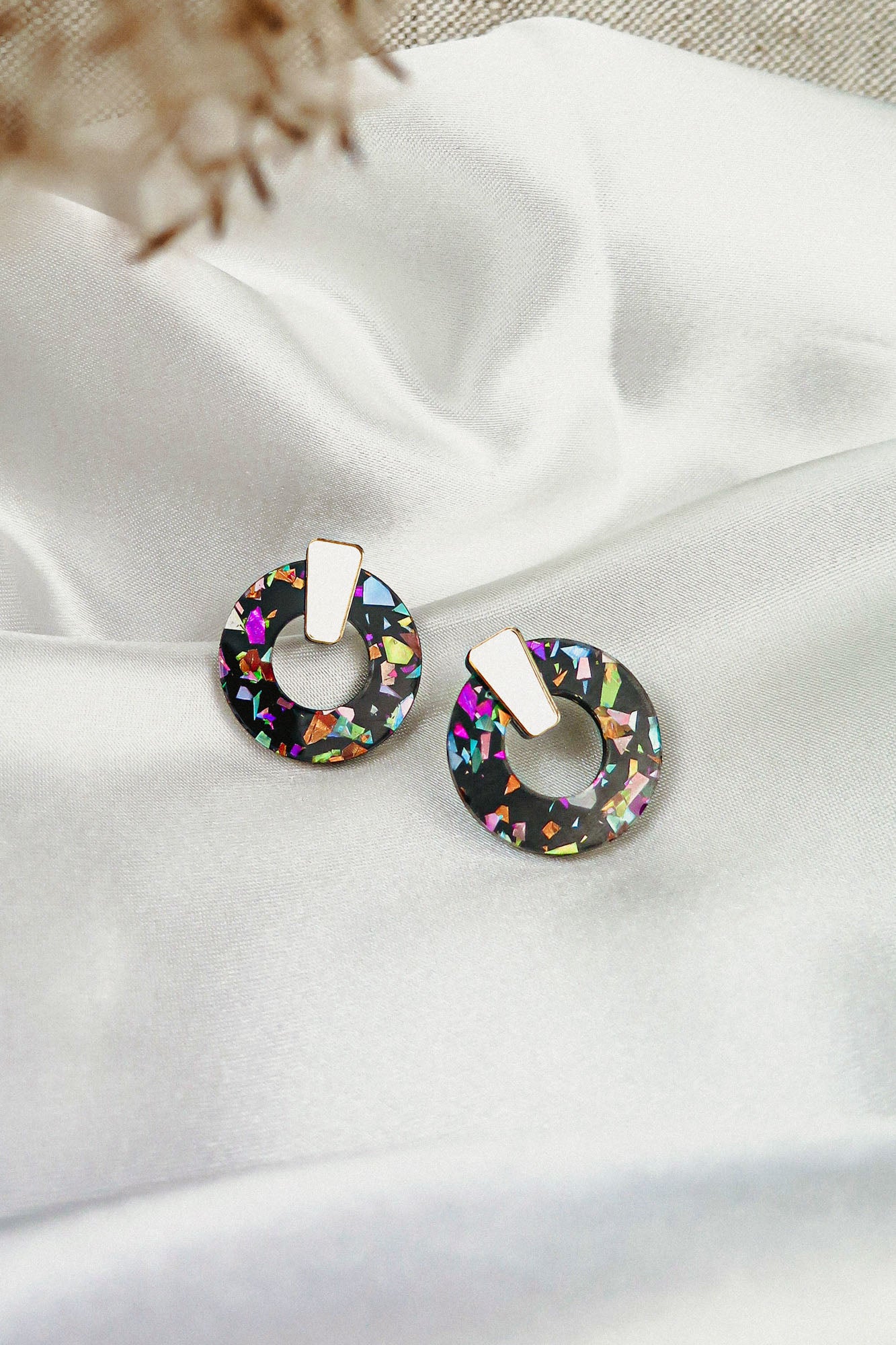 A pair of round, black Minimalist Circle Stud Earrings with colorful iridescent flecks and gold rectangular studs, crafted from hypoallergenic acrylic glass, displayed on white satin fabric.