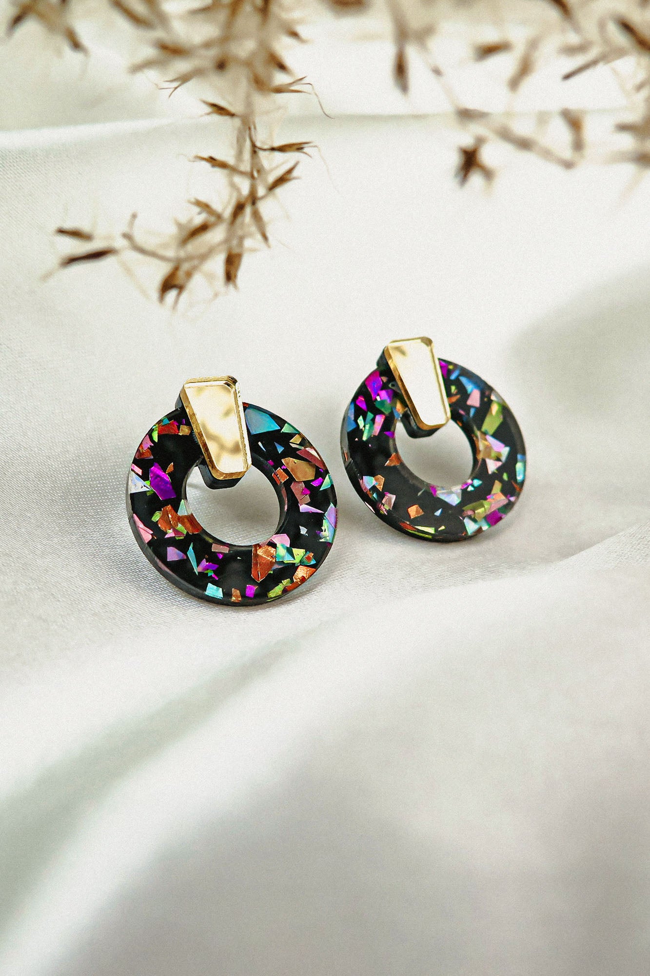 Close-up of Minimalist Circle Stud Earrings with gold tops and black acrylic glass bases featuring colorful, iridescent mosaic fragments, displayed on a white fabric background with dried plant stems overhead. Hypoallergenic design.