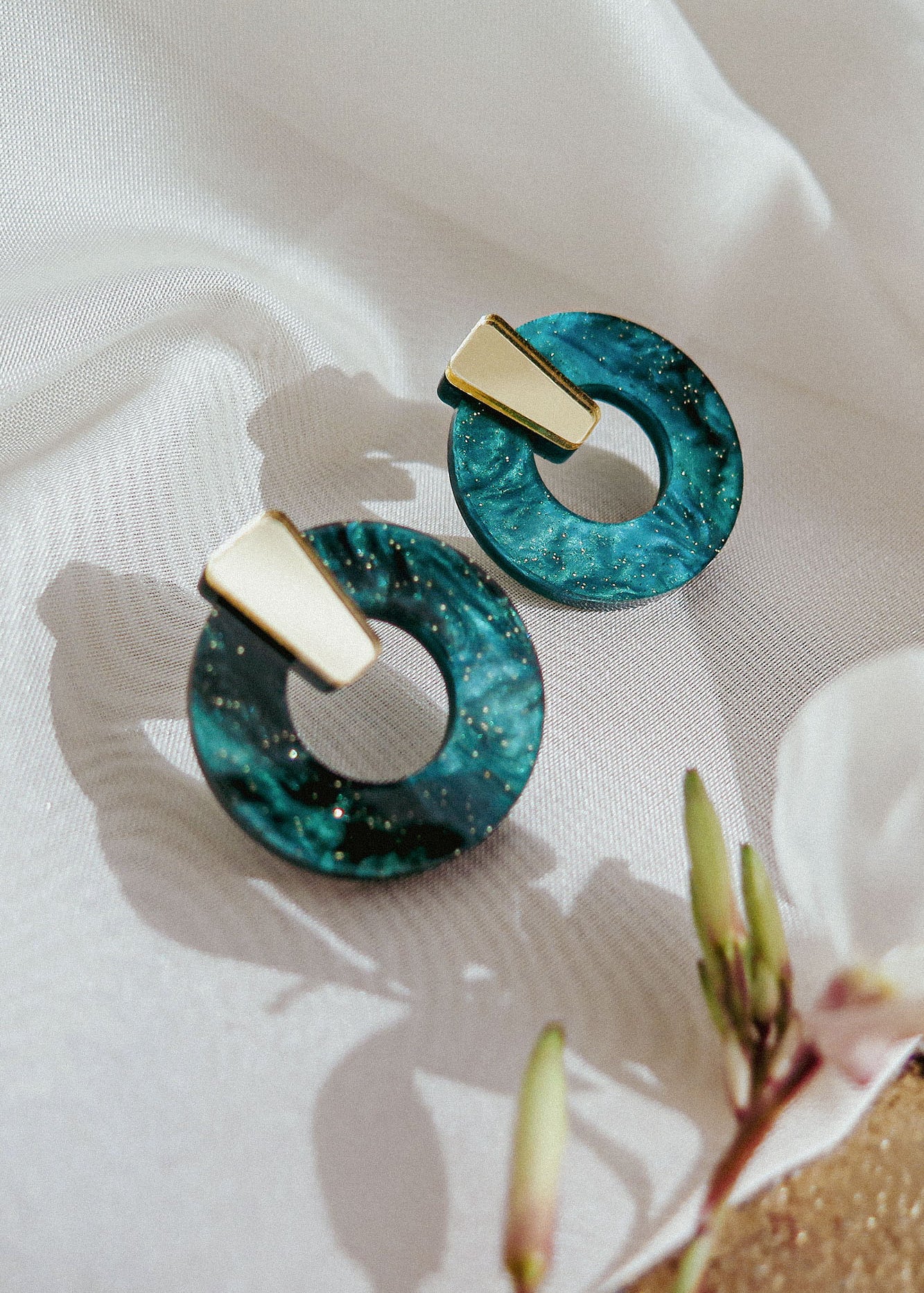 A pair of hypoallergenic, modern earrings with gold rectangular tops and circular blue-green marbled resin hoops, displayed on white fabric with a flower and buds nearby.