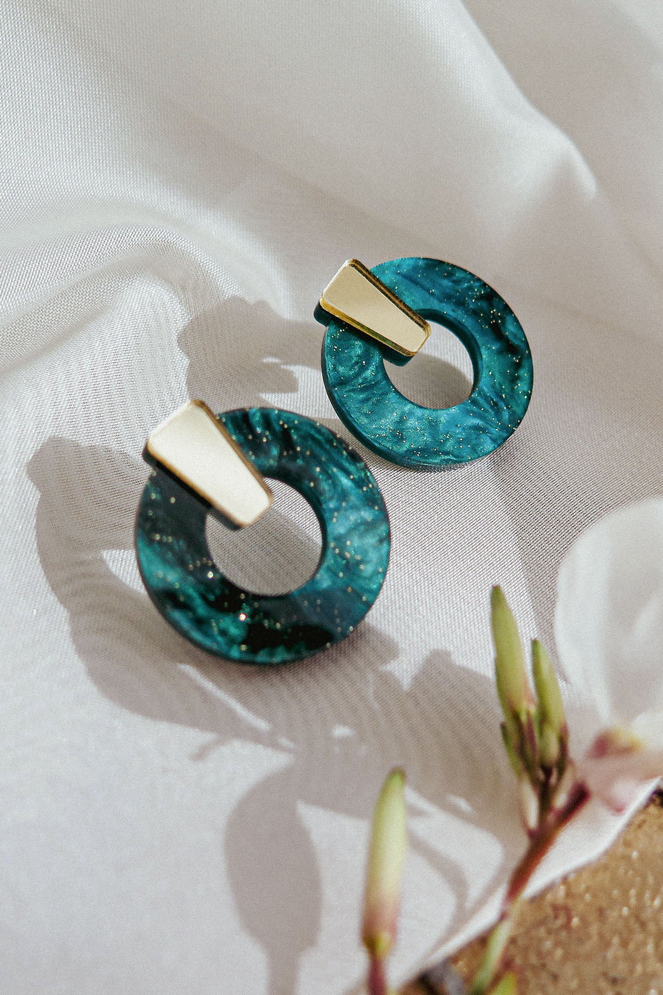 A pair of hypoallergenic, modern earrings with gold rectangular tops and circular blue-green marbled resin hoops, displayed on white fabric with a flower and buds nearby.