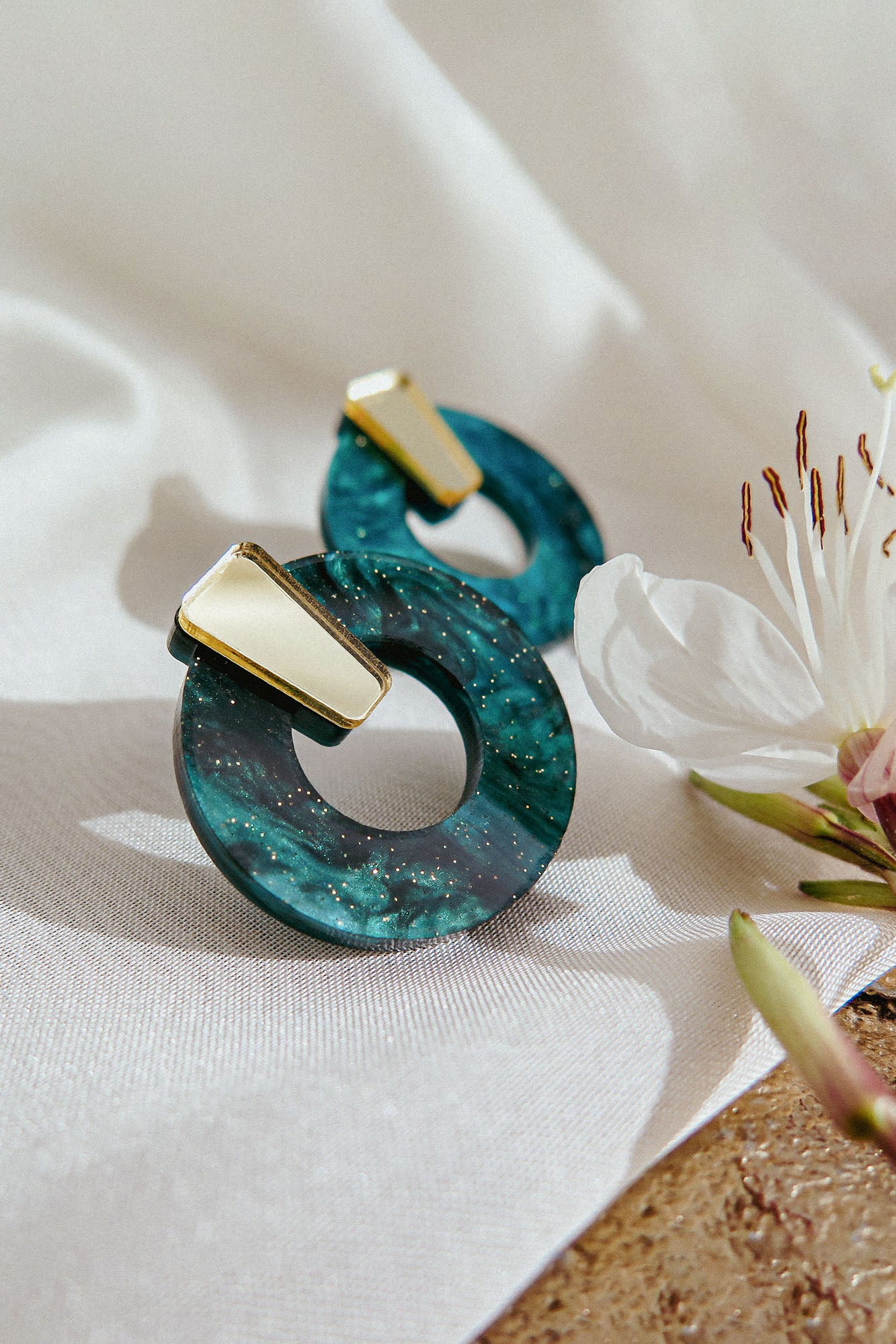 Two minimalist circle stud earrings with a marbled teal design and small gold accent, crafted from hypoallergenic acrylic glass, are displayed on a white cloth next to a white flower with pink details.