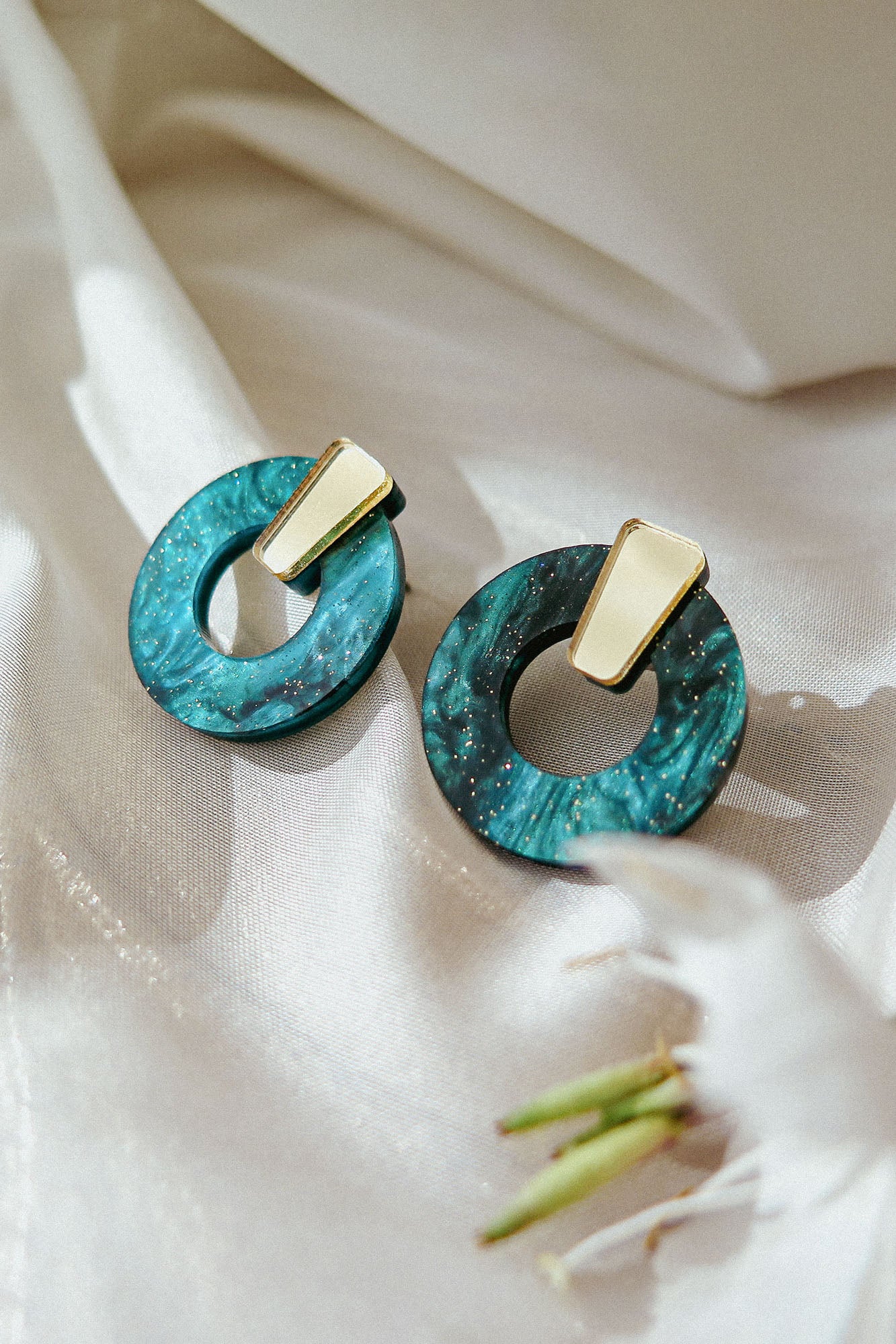 A pair of minimalist circle stud earrings crafted from teal marbled acrylic glass with gold geometric accents rest on shiny white fabric, with a blurred white flower in the foreground. Hypoallergenic for sensitive ears.