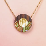A minimalistic gold chain Circle Necklace with an oval pendant featuring wood, iridescent abalone shell, and a central circular gold detail, perfect for elevating any capsule wardrobe. Displayed against a pale pink background.