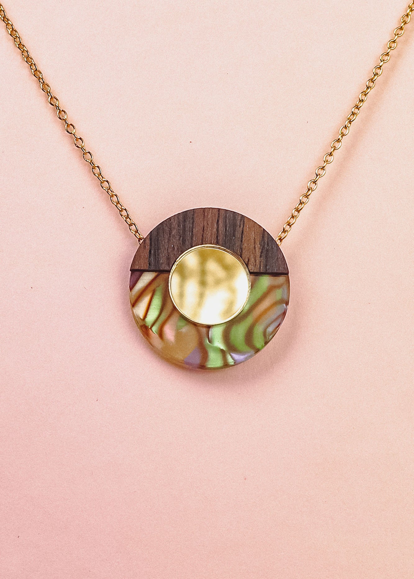 A minimalistic gold chain Circle Necklace with an oval pendant featuring wood, iridescent abalone shell, and a central circular gold detail, perfect for elevating any capsule wardrobe. Displayed against a pale pink background.