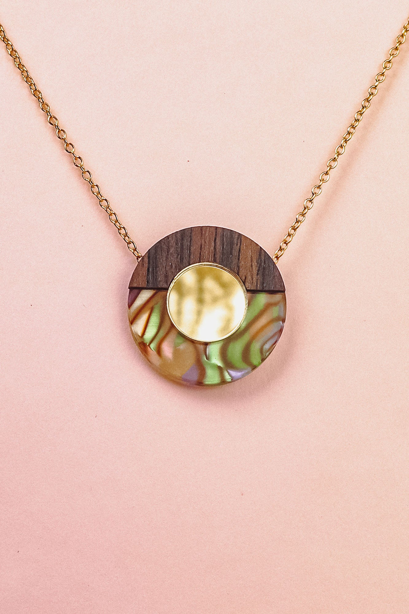 A minimalistic gold chain Circle Necklace with an oval pendant featuring wood, iridescent abalone shell, and a central circular gold detail, perfect for elevating any capsule wardrobe. Displayed against a pale pink background.