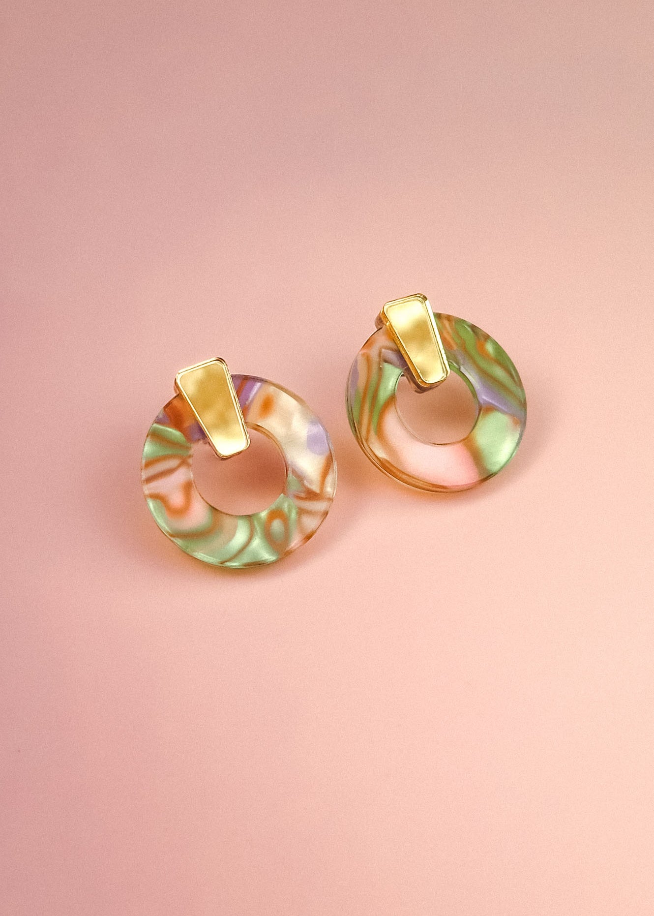 A pair of minimalist circle stud earrings crafted from acrylic glass in marbled pastel green, pink, and purple, with gold hypoallergenic rectangular studs, displayed on a soft pink background.