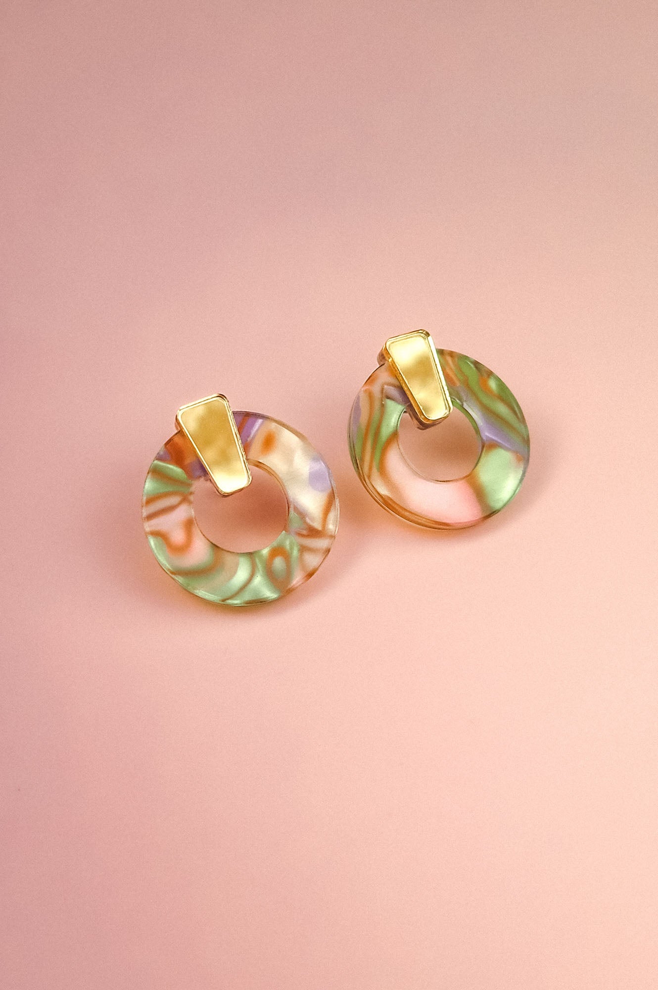 A pair of minimalist circle stud earrings crafted from acrylic glass in marbled pastel green, pink, and purple, with gold hypoallergenic rectangular studs, displayed on a soft pink background.