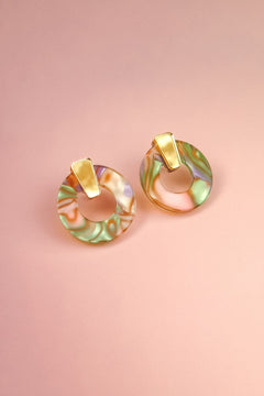 A pair of minimalist circle stud earrings crafted from acrylic glass in marbled pastel green, pink, and purple, with gold hypoallergenic rectangular studs, displayed on a soft pink background.