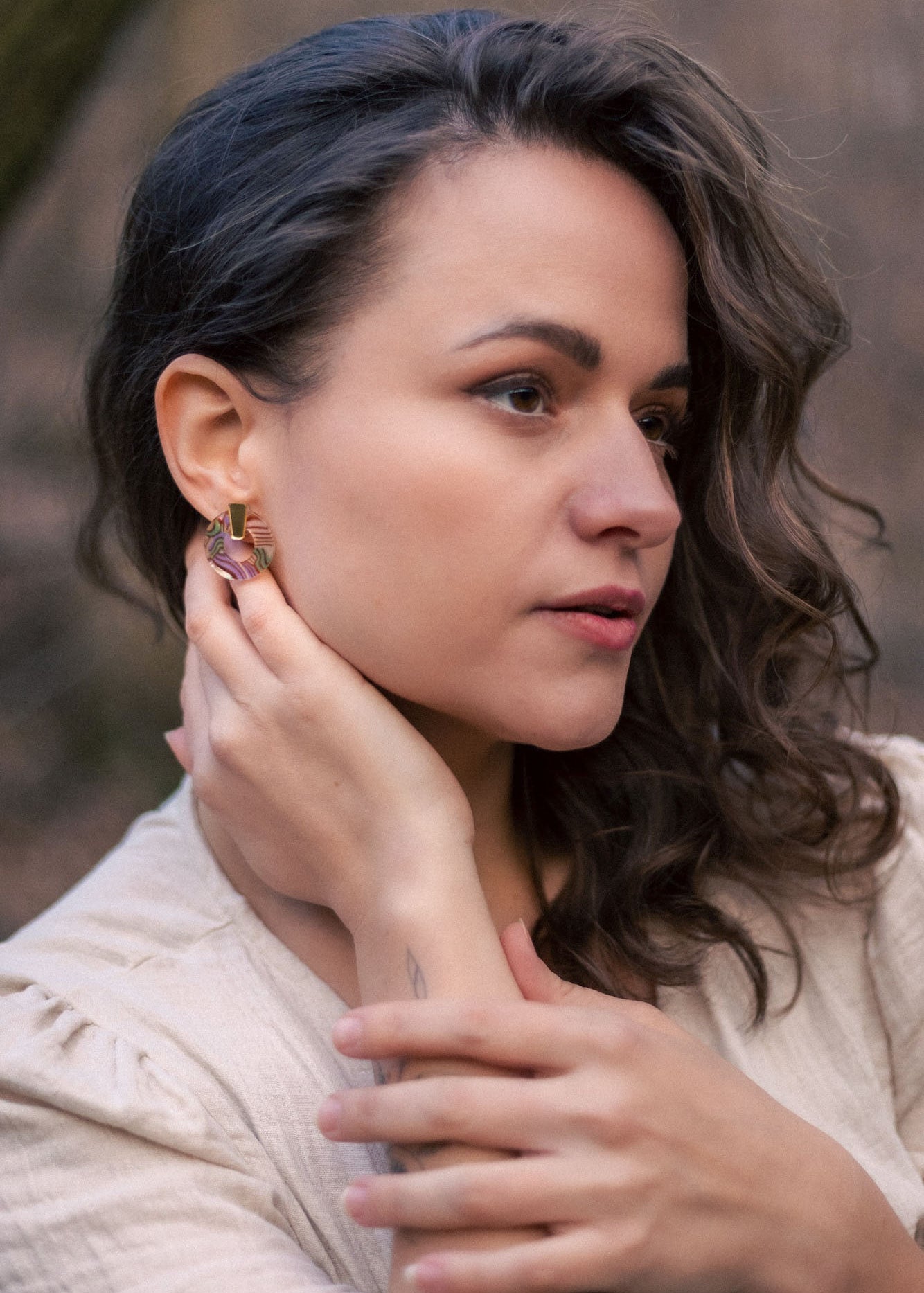 A woman with wavy brown hair wearing a beige top touches her Minimalist Circle Stud Earrings with one hand and rests her other hand on her collarbone. She looks slightly to the side with a calm expression. The earrings are hypoallergenic.