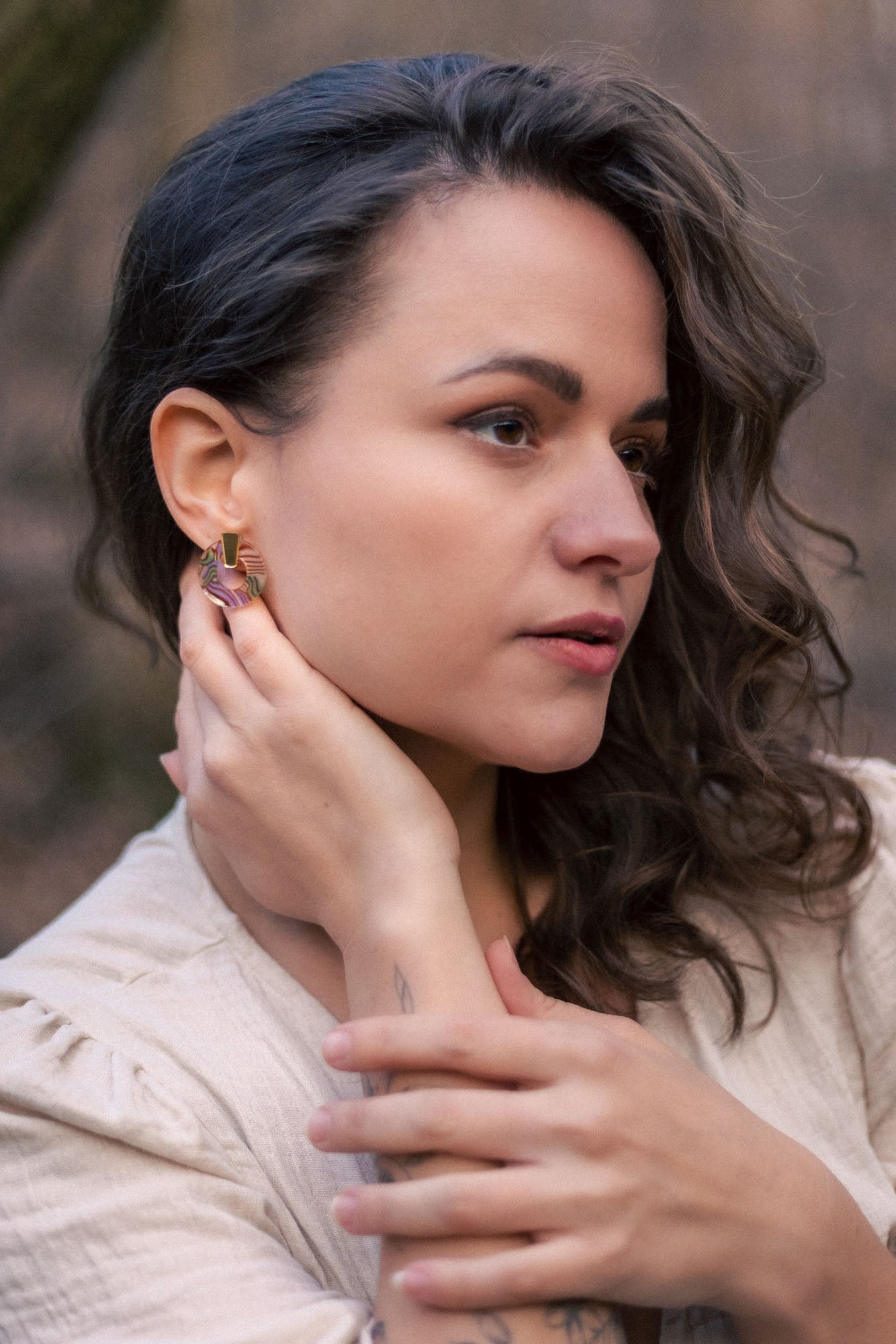 A woman with wavy brown hair wearing a beige top touches her Minimalist Circle Stud Earrings with one hand and rests her other hand on her collarbone. She looks slightly to the side with a calm expression. The earrings are hypoallergenic.