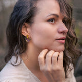 A woman with wavy brown hair and a light beige blouse looks to the side outdoors, resting her chin on her hand. She wears colorful hypoallergenic Minimalist Circle Stud Earrings and has a subtle tattoo on her wrist.