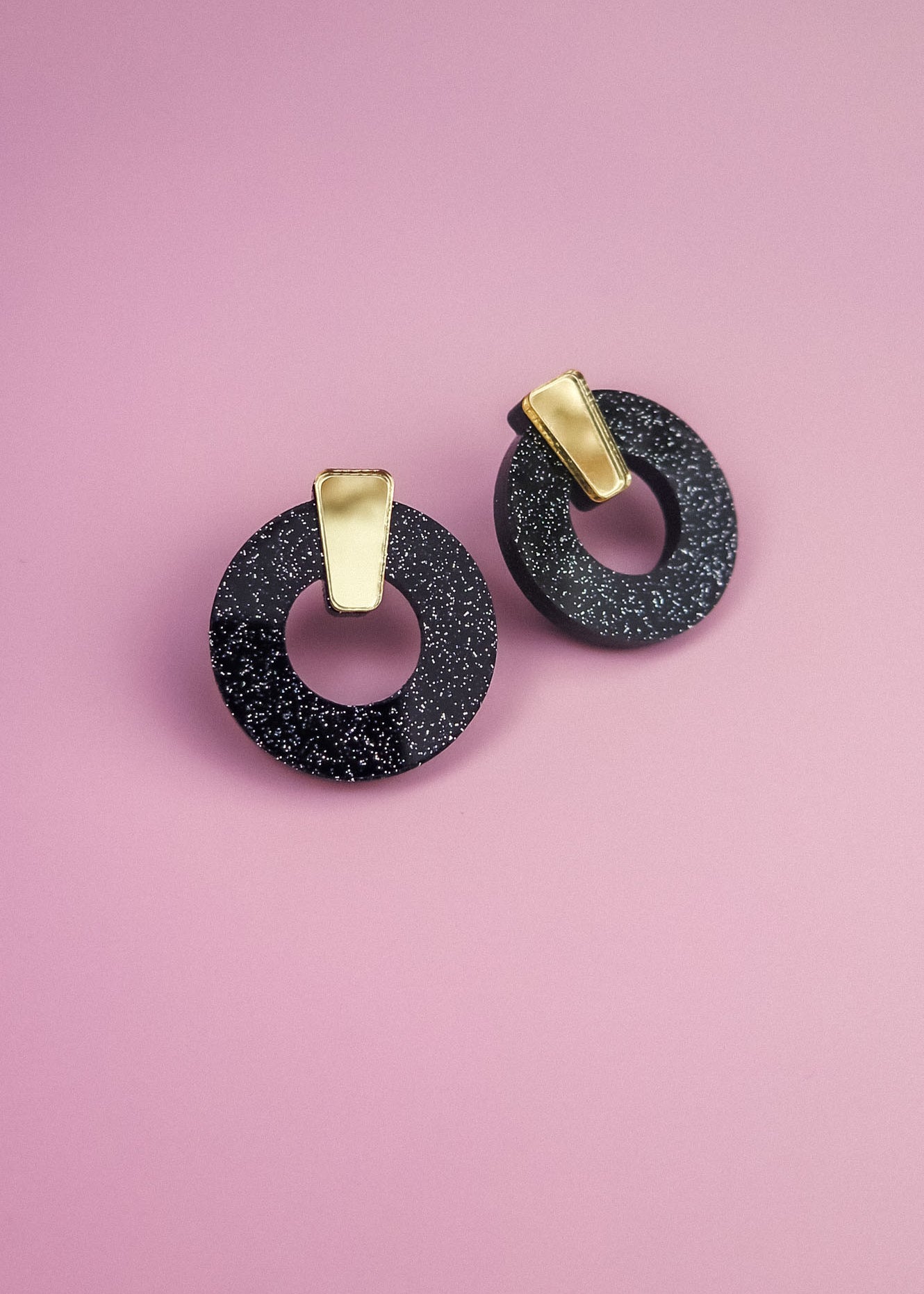 A pair of minimalist circle stud earrings, crafted from hypoallergenic acrylic glass with white speckles and gold rectangular accents, are displayed on a pink background.