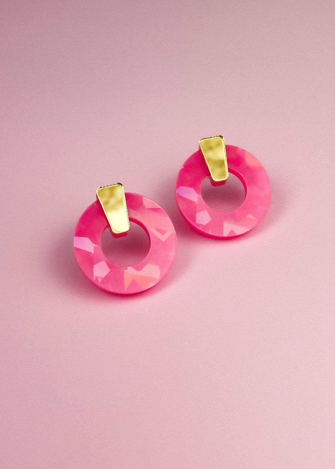 A pair of Minimalist Circle Stud Earrings featuring circular, faceted pink acrylic glass with elegant gold accents, displayed on a soft pink background.