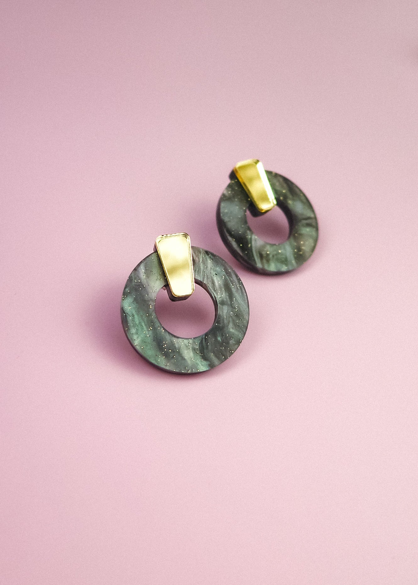 A pair of hypoallergenic Minimalist Circle Stud Earrings, featuring round green marbled acrylic glass and gold rectangular studs, displayed on a pink background.