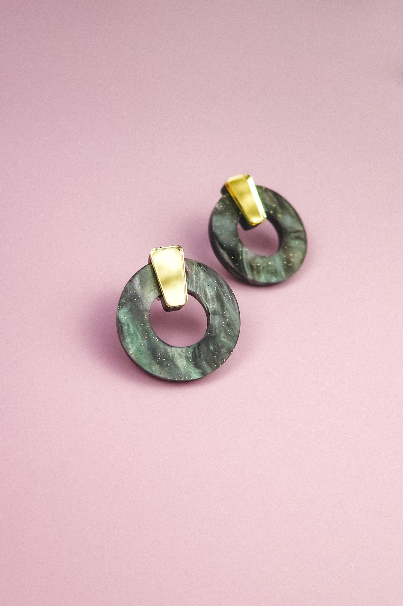 A pair of hypoallergenic Minimalist Circle Stud Earrings, featuring round green marbled acrylic glass and gold rectangular studs, displayed on a pink background.