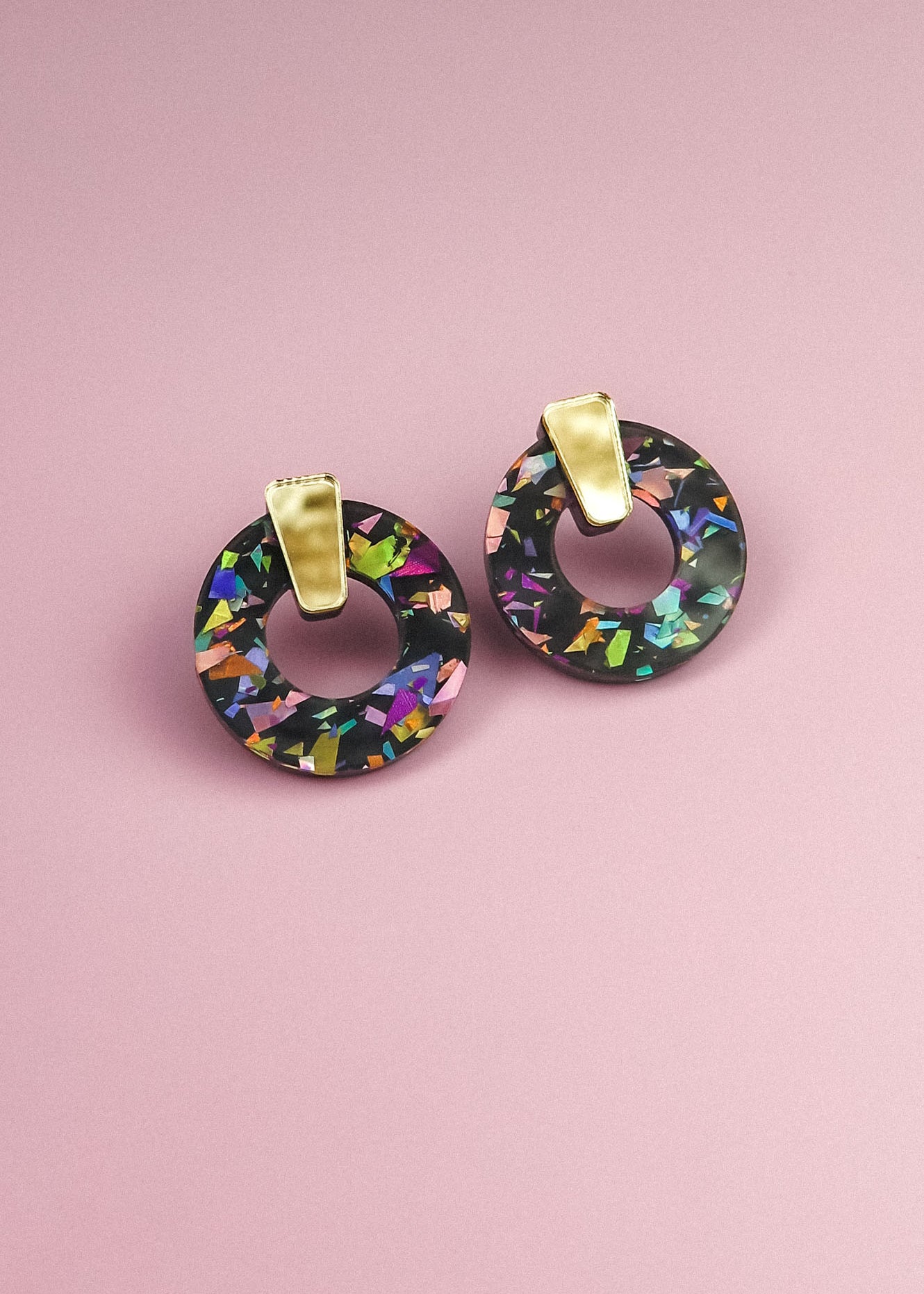 A pair of minimalist circle stud earrings with multicolored, iridescent patterns crafted from acrylic glass and gold rectangular clips, displayed on a pink background.
