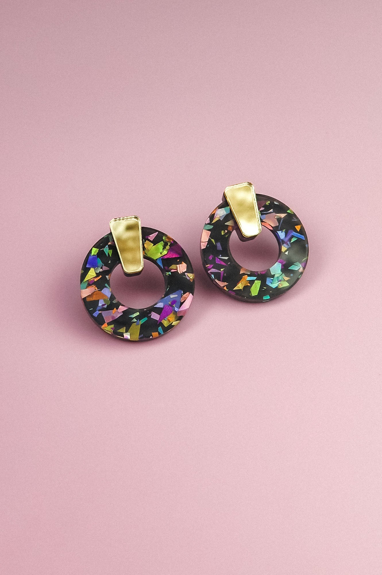 A pair of minimalist circle stud earrings with multicolored, iridescent patterns crafted from acrylic glass and gold rectangular clips, displayed on a pink background.