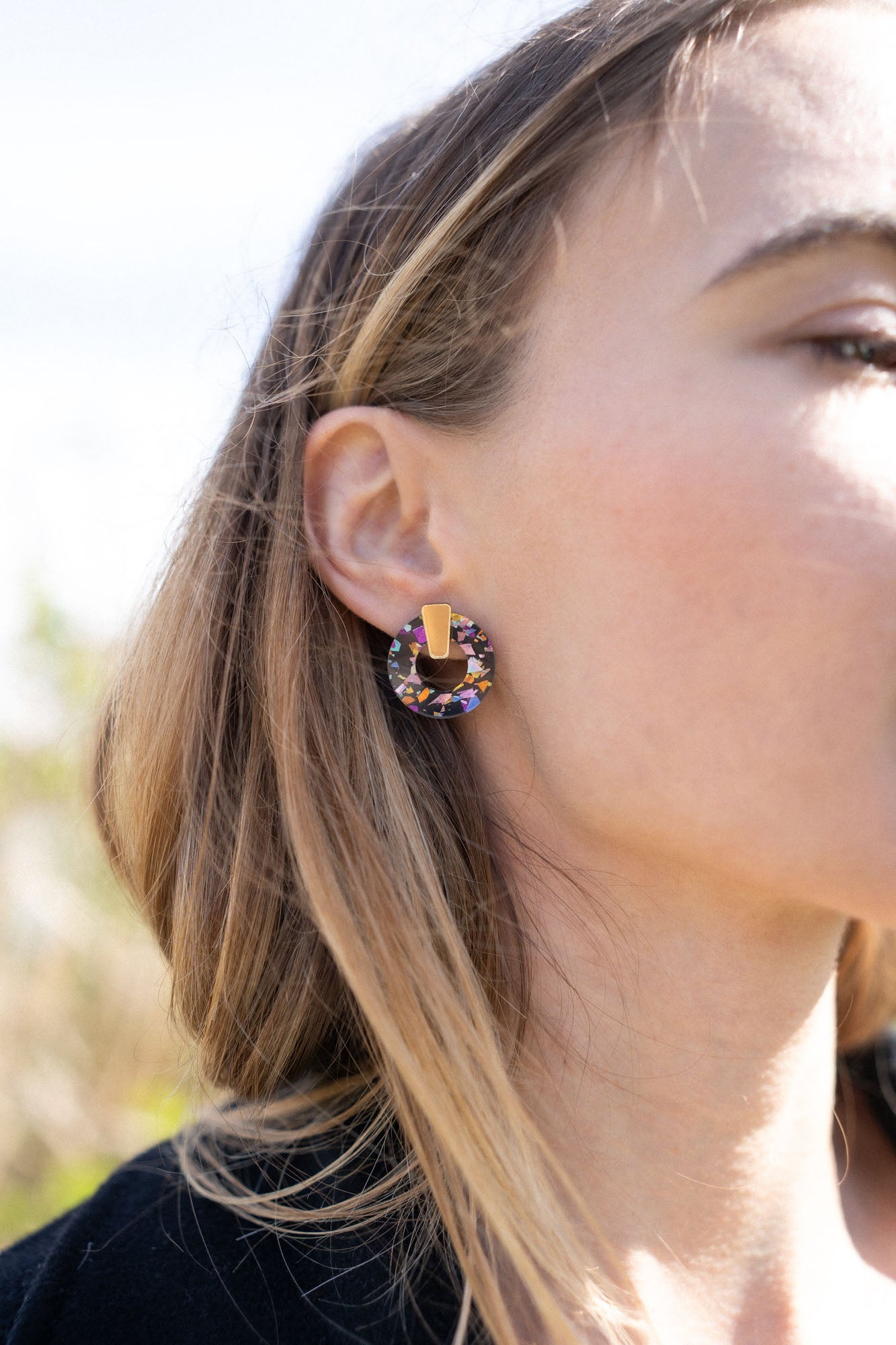 A close-up of a woman wearing Minimalist Circle Stud Earrings crafted from hypoallergenic materials, featuring a large, circular, multicolored gemstone with a gold accent, shown from the side against a blurred outdoor background.