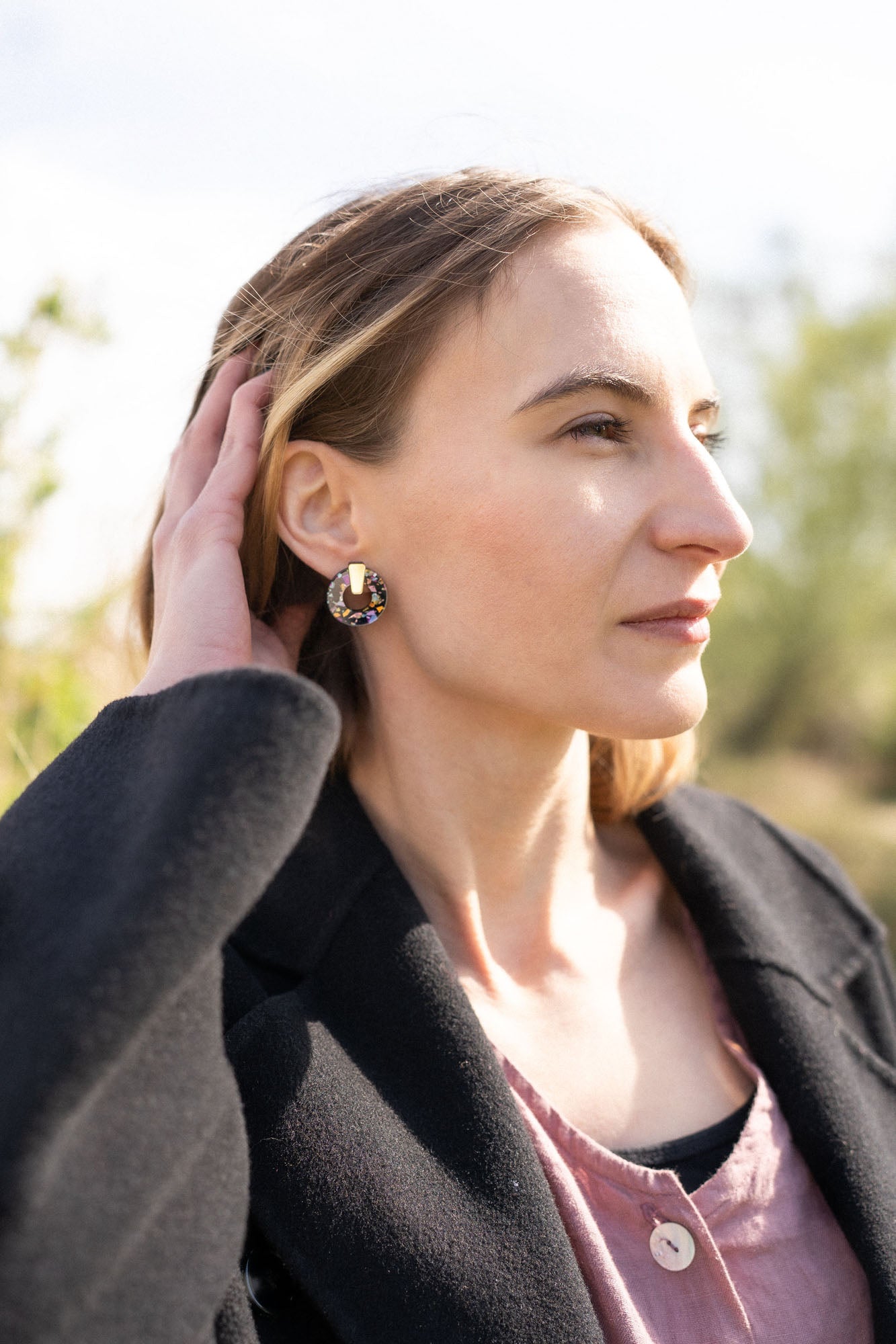 A woman with light brown hair touches her head while looking into the distance. She wears a black coat, a mauve top, and Minimalist Circle Stud Earrings that are hypoallergenic. The background is outdoors and softly blurred by sunlight.